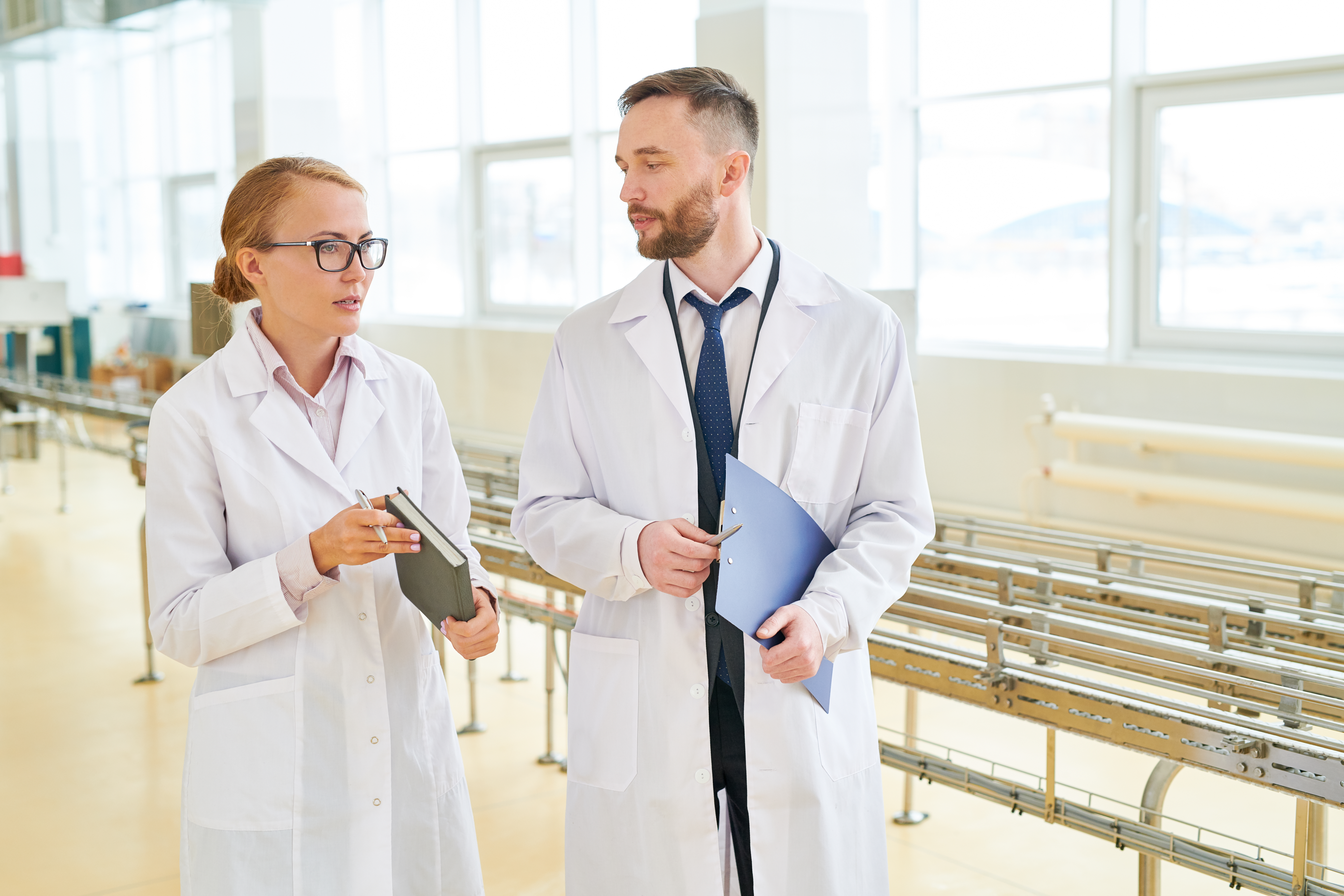 Beautiful technologist wearing white coat having small talk with bearded inspector while giving tour of production department of milk plant